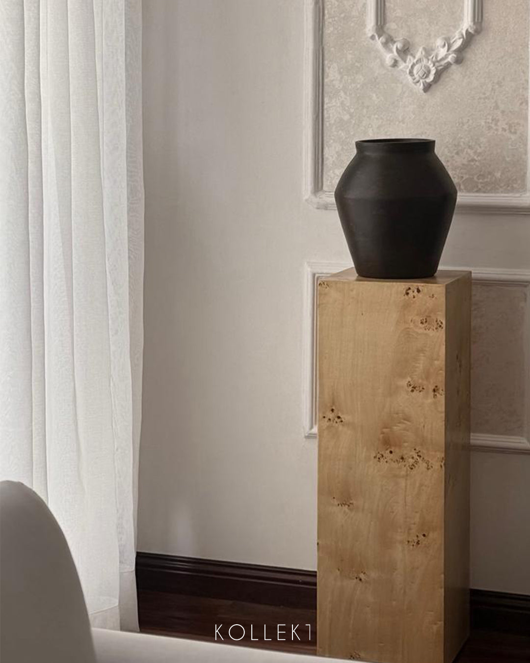 Black vase on a wooden pedestal against a neutral wall with a mirror.