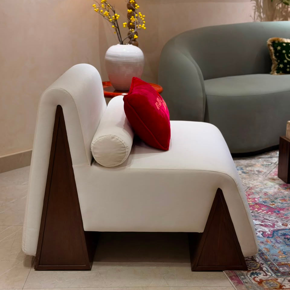 A modern white upholstered lounge chair with dark brown geometric wooden legs, styled with a white cylindrical pillow and a red accent pillow in a living room setting.